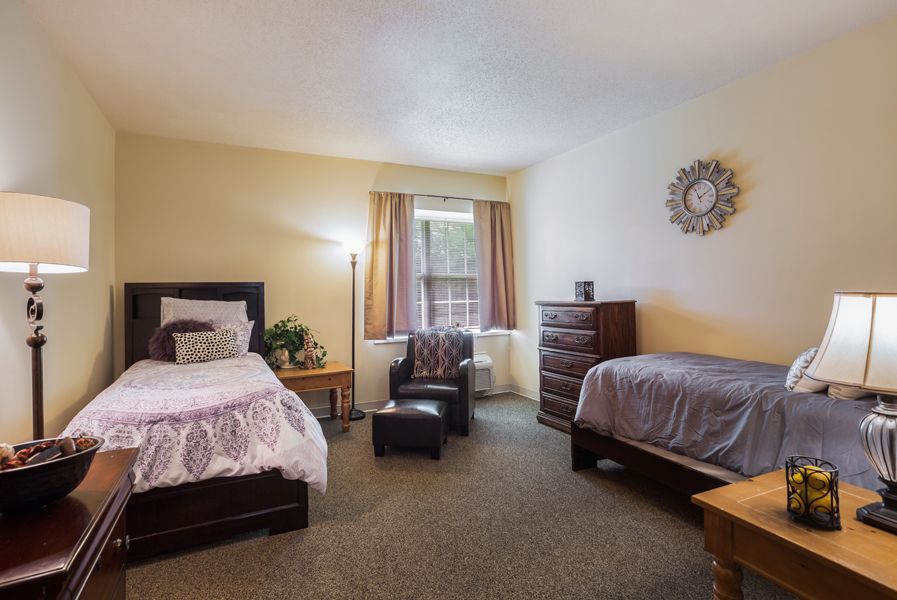 A cozy bedroom with two beds, a chair, and a dresser, featuring warm lighting and a window.