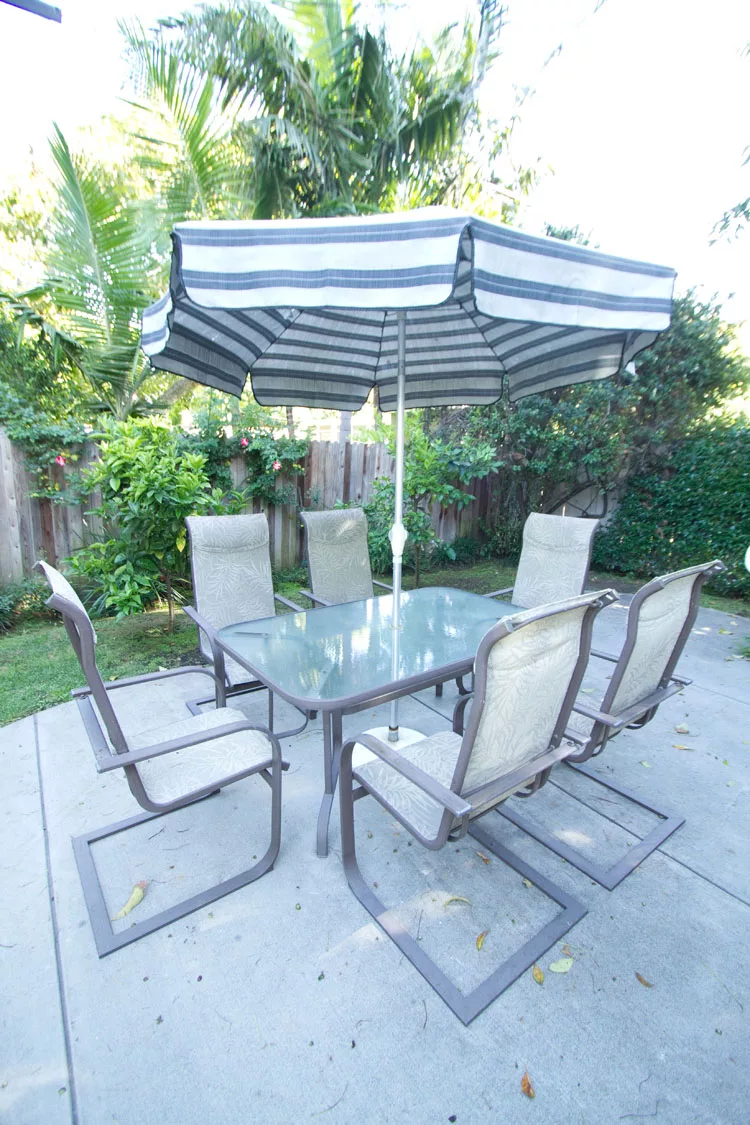 A glass patio table with six chairs and a striped umbrella, set in a garden.