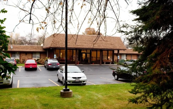 A small commercial building with a parking lot, surrounded by trees under an overcast sky.