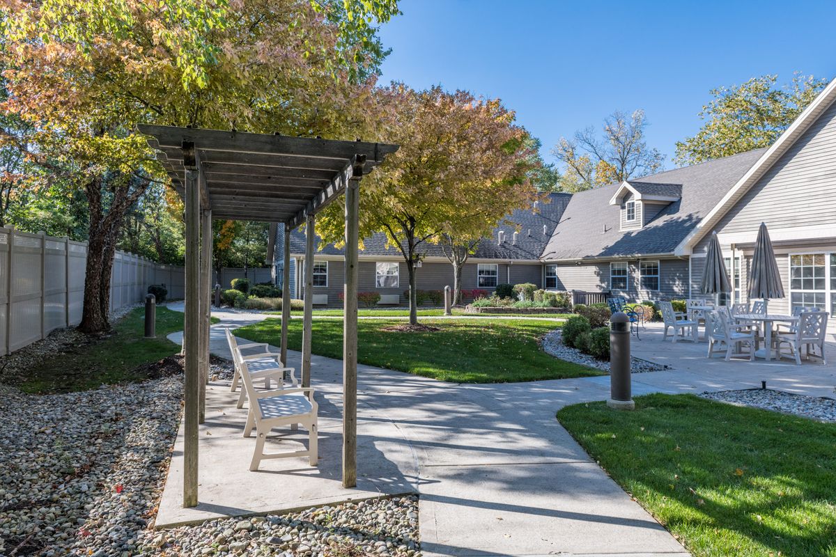 A peaceful garden pathway with benches under a pergola, surrounded by trees and houses on a sunny day.