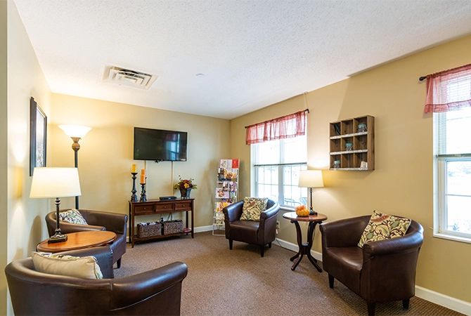 A cozy living room with comfortable chairs, a table, a mounted television, and a large window allowing natural light to enter.