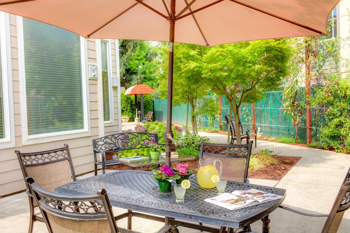 A cozy outdoor patio setting with a table, chairs, and an umbrella surrounded by lush greenery.