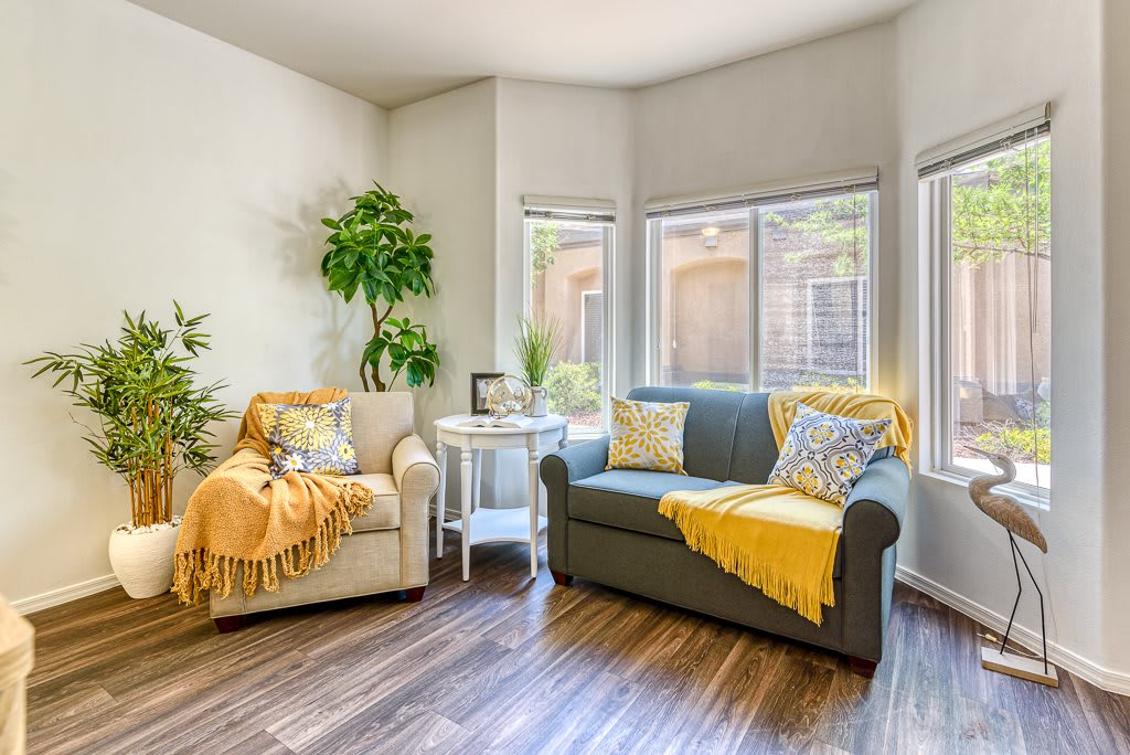A cozy living room with a sofa and armchair, accented by yellow throws and greenery, filled with natural light from large windows.