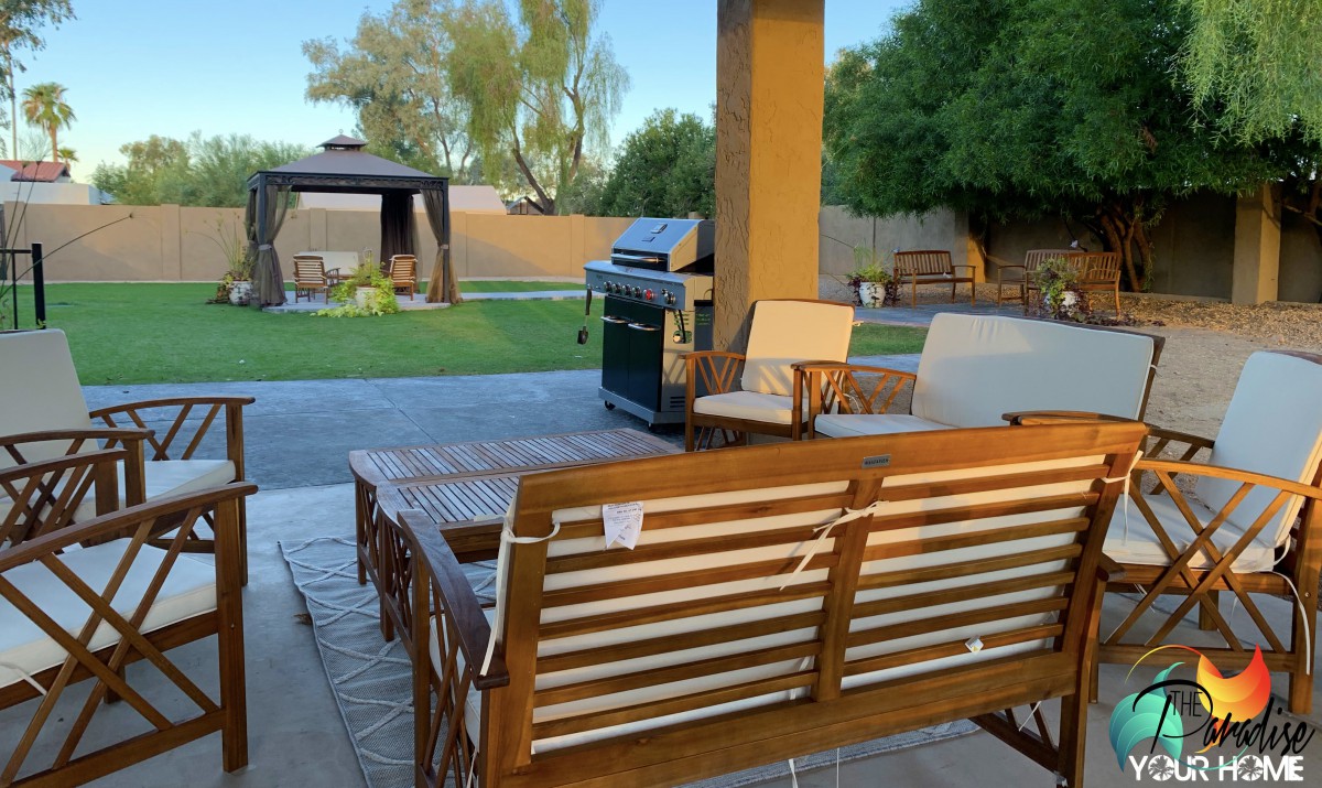 A cozy backyard patio with wooden furniture and a grill, overlooking a lawn and gazebo.
