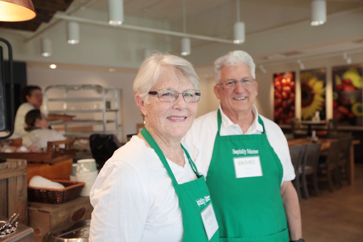A senior couple volunteering in a kitchen, wearing green aprons and smiling.