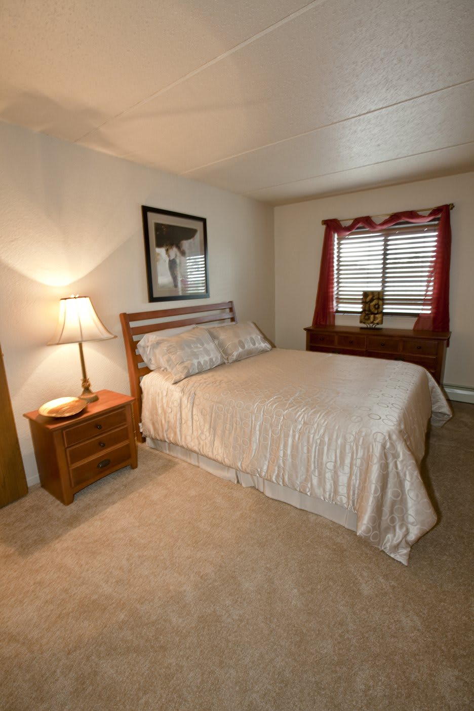 A cozy bedroom with a neatly made bed, nightstand, and lamp next to a window with red curtains.