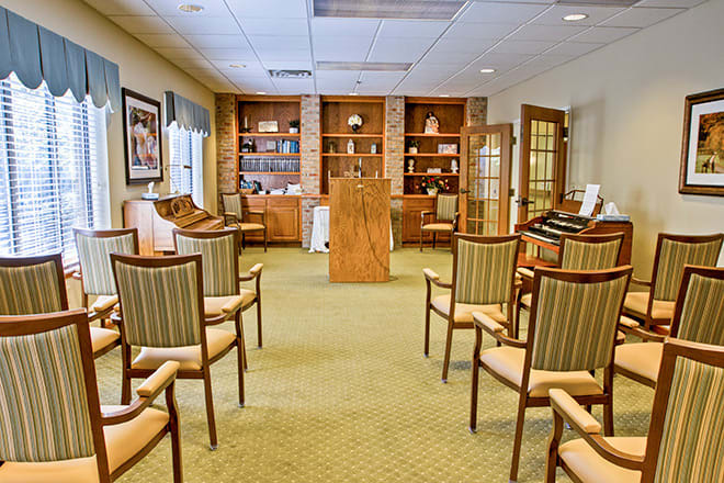 An indoor chapel with arranged chairs, a central podium, and bookshelves in the background.
