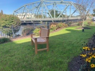 A peaceful outdoor scene featuring a wooden bench on green grass with a bridge over a river in the background.