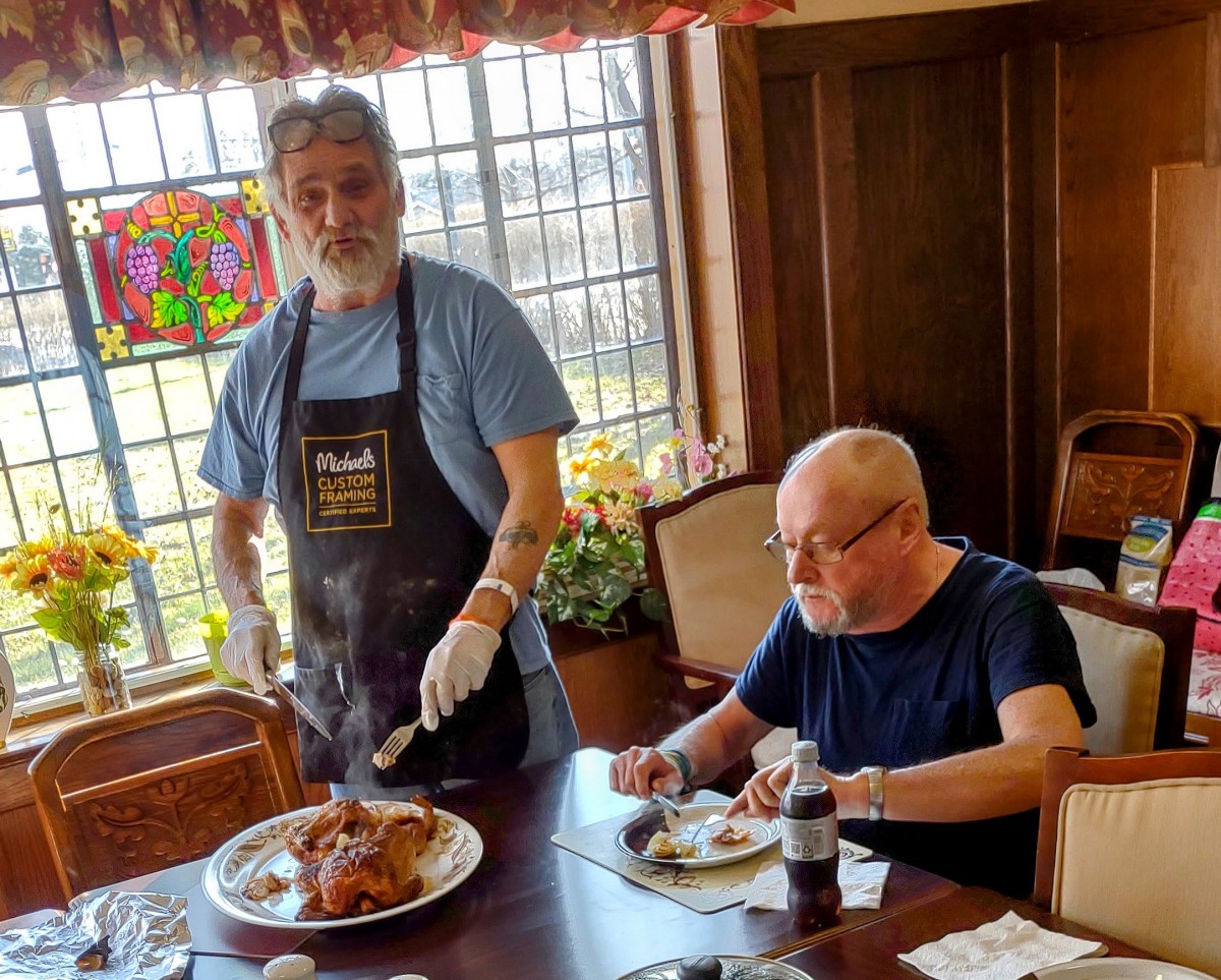 Two individuals are at a dining table; one is seated enjoying a meal while the other stands nearby, preparing to serve food.