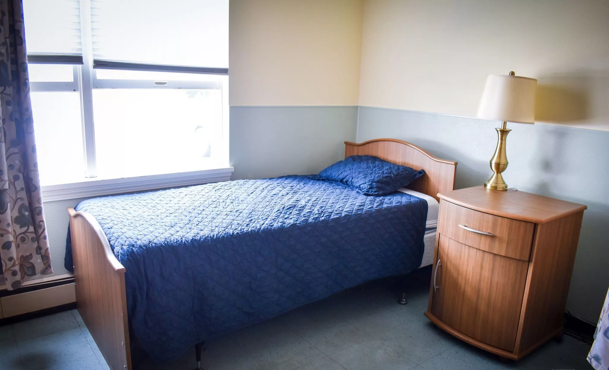 A simple bedroom with a single bed and blue bedding next to a window.