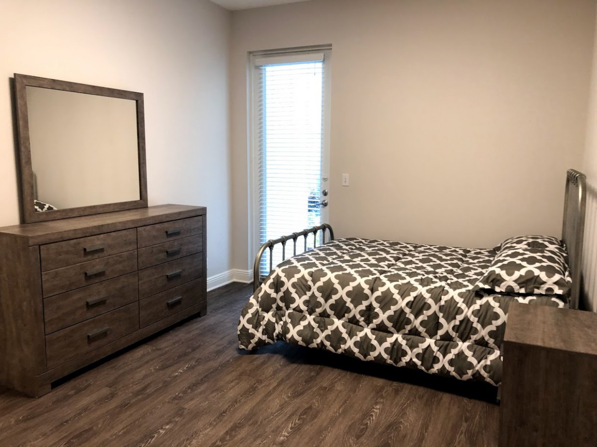 A modern bedroom featuring a neatly made bed with patterned bedding, a wooden dresser with a mirror, and a wooden floor.