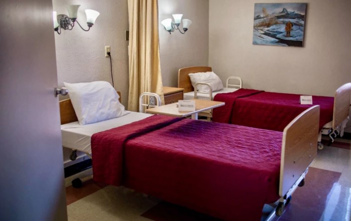 A hospital room with two neatly arranged twin beds, featuring red blankets and bedside tables.