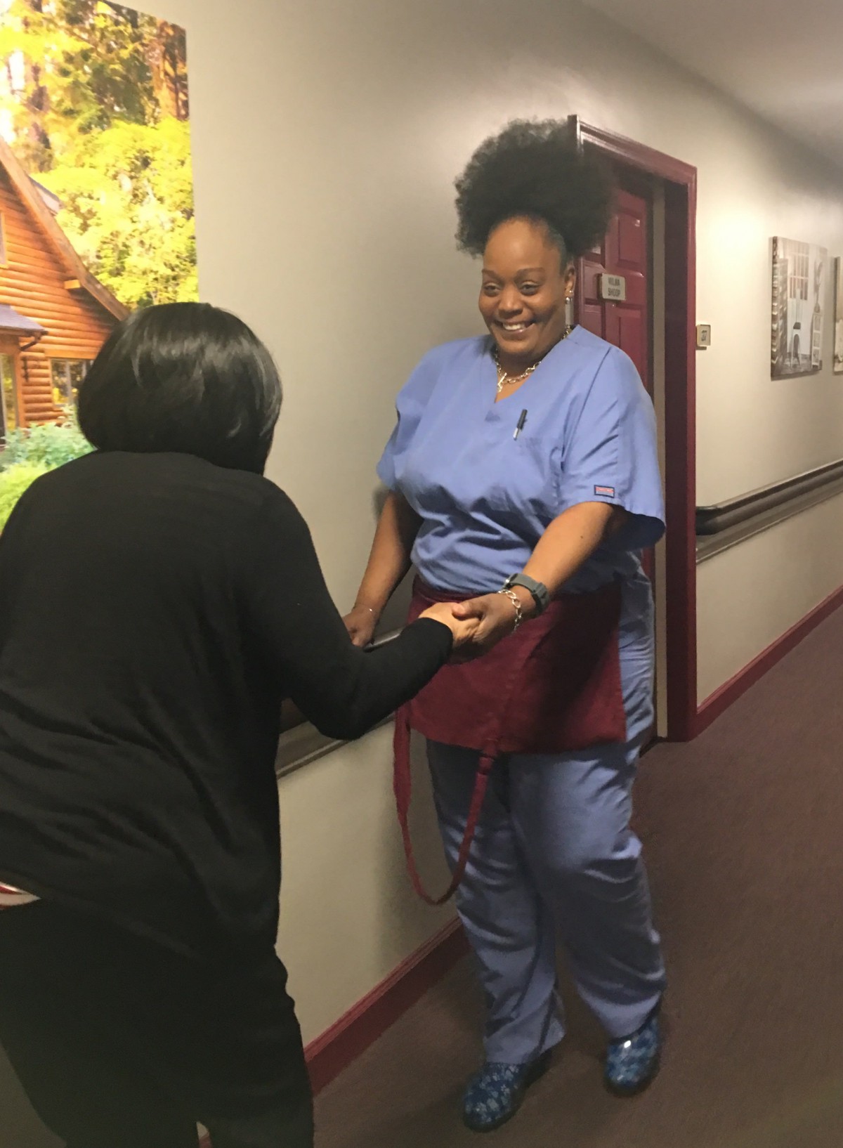 A nurse in blue scrubs smiles while interacting with another person in a hallway.
