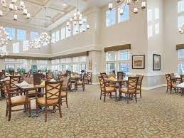A spacious and well-lit dining area with multiple tables and chairs, featuring large windows and elegant chandeliers.