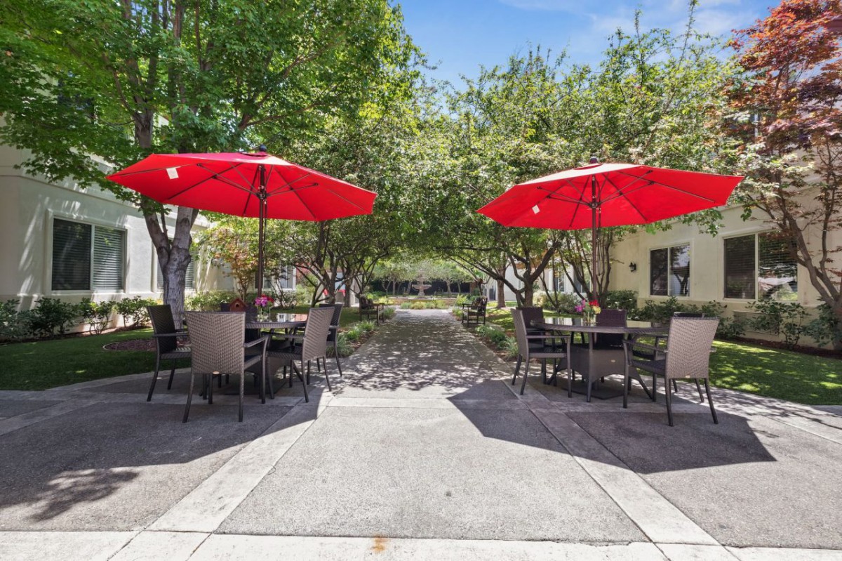A cozy outdoor patio area with tables, chairs, and red umbrellas surrounded by greenery.