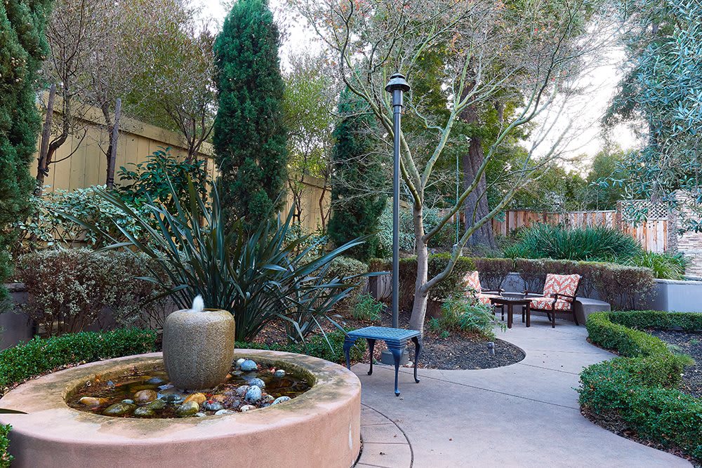 A serene outdoor garden area featuring a small fountain, surrounded by lush greenery and comfortable patio seating.