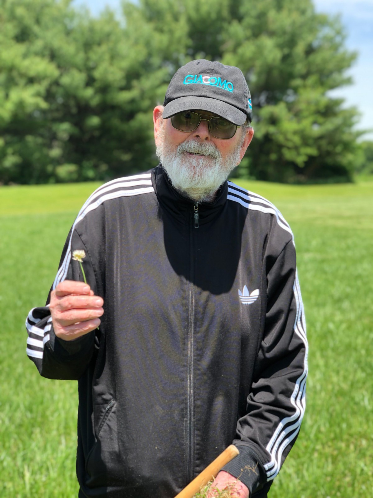 A bearded man in a black jacket and hat standing outside on a grassy field.