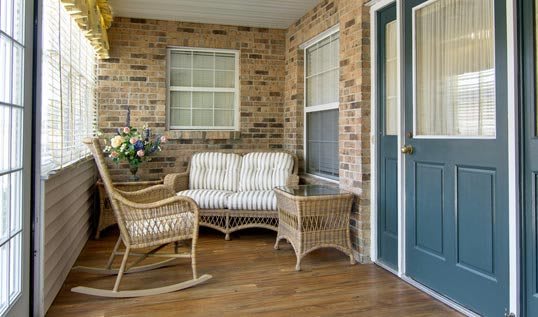 A cozy porch with wicker furniture including a sofa, chairs, and a small table, set against a brick wall.