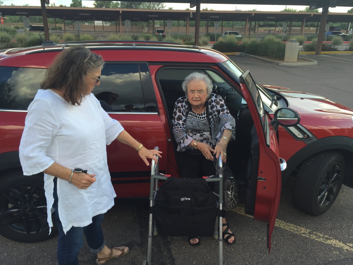 An elderly woman is assisted by another woman as she exits a car using a walker in a parking lot.