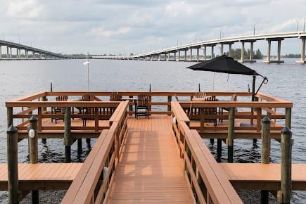 A dock with outdoor seating and an umbrella overlooks a river with a bridge in the background.