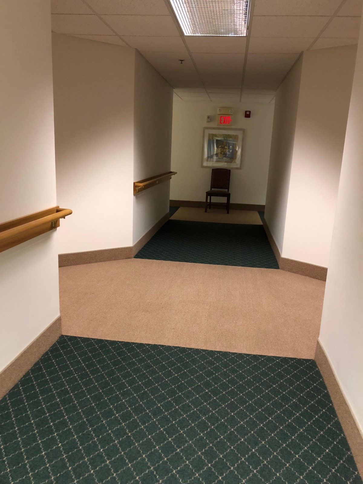 A carpeted hallway with handrails on both sides and a chair near an exit sign at the end.