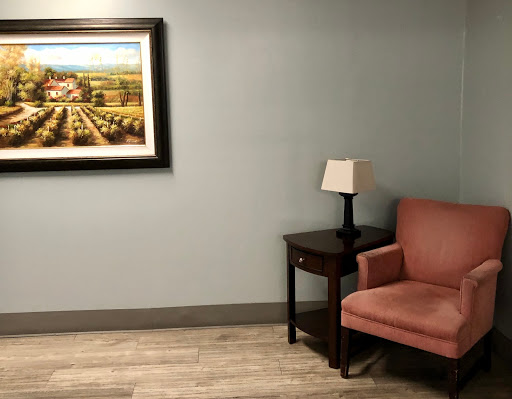 A cozy lounge area featuring a pink chair, a small table with a lamp, and framed artwork on the wall.