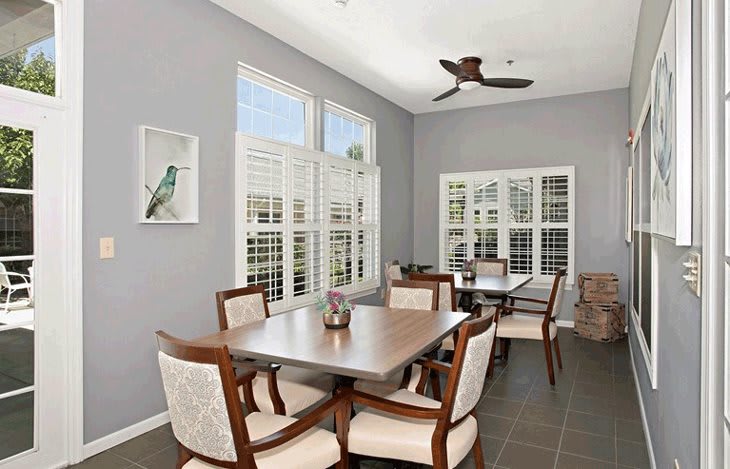 A well-lit dining room featuring a wooden table with six chairs, large windows with shutters, and a ceiling fan.