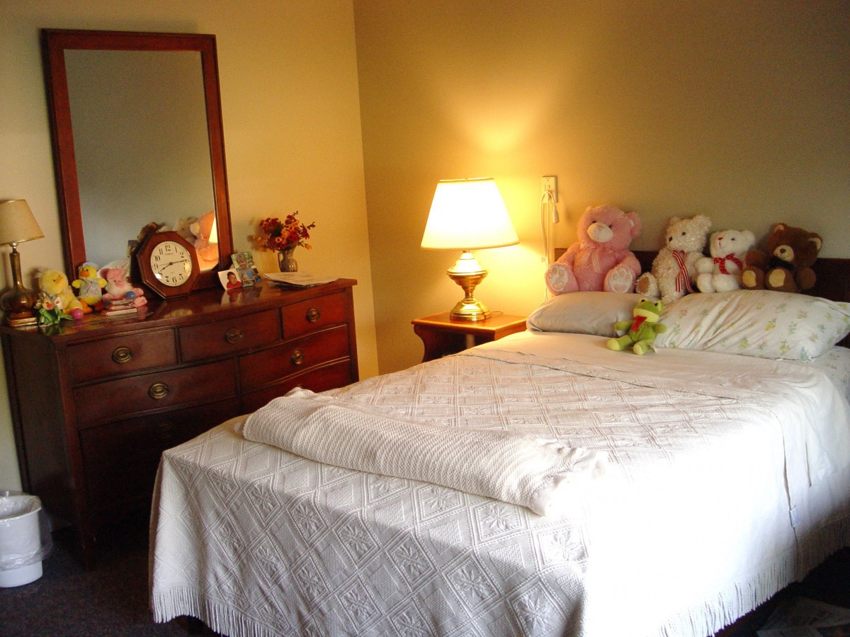 A cozy bedroom with a neatly made bed, soft lighting from a lamp, and stuffed animals arranged on the pillows.