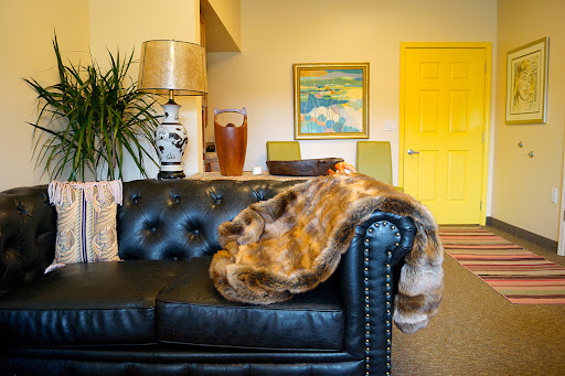A stylish living room featuring a black leather sofa draped with a faux fur throw, accompanied by colorful artwork and a vibrant yellow door.