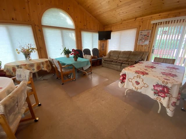 A cozy living and dining area with wood paneling, featuring a sofa, tables, chairs, and a television. Large windows let in ample sunlight.