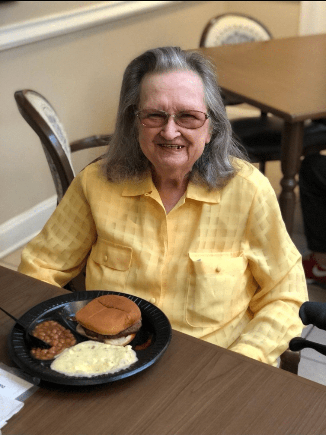 An elderly woman smiling while sitting in front of a meal at a table.