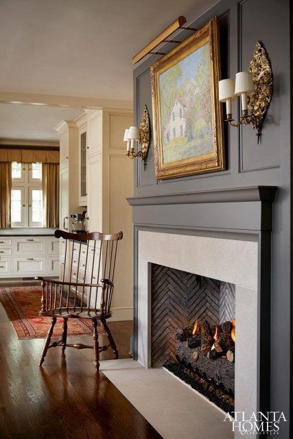 A cozy living room featuring a fireplace with a mounted painting, a wooden rocking chair, and elegant wall sconces.