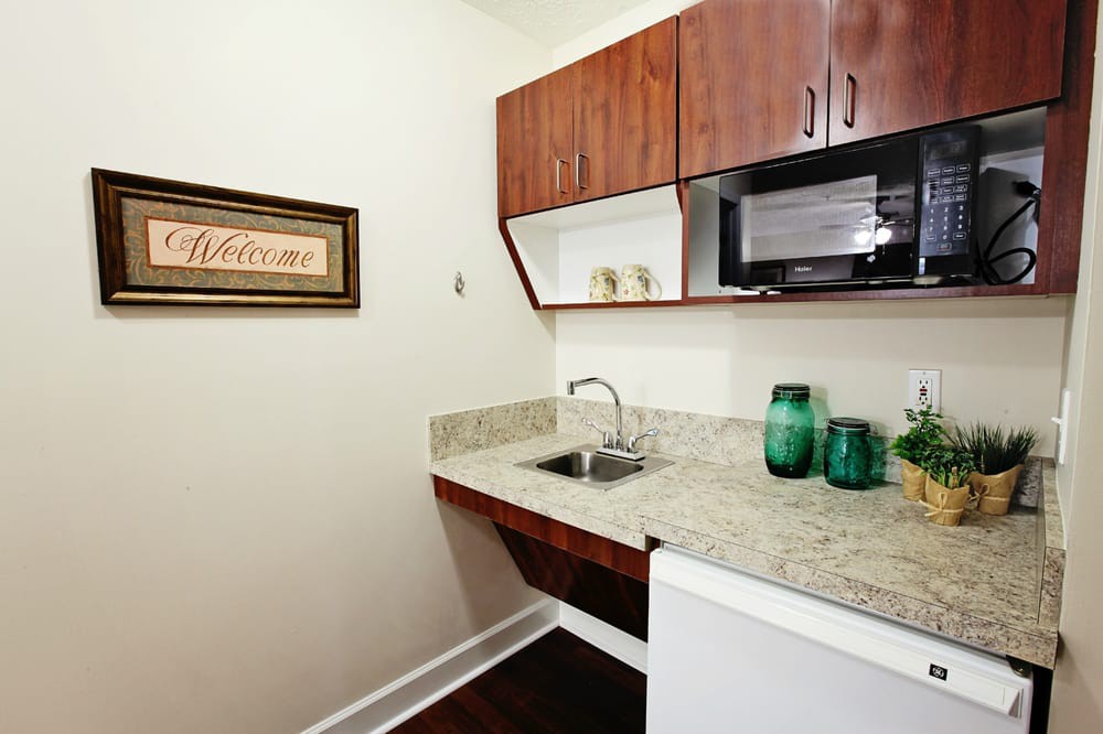 A small kitchenette with wooden cabinets, granite countertop, a microwave, and decorative accents.