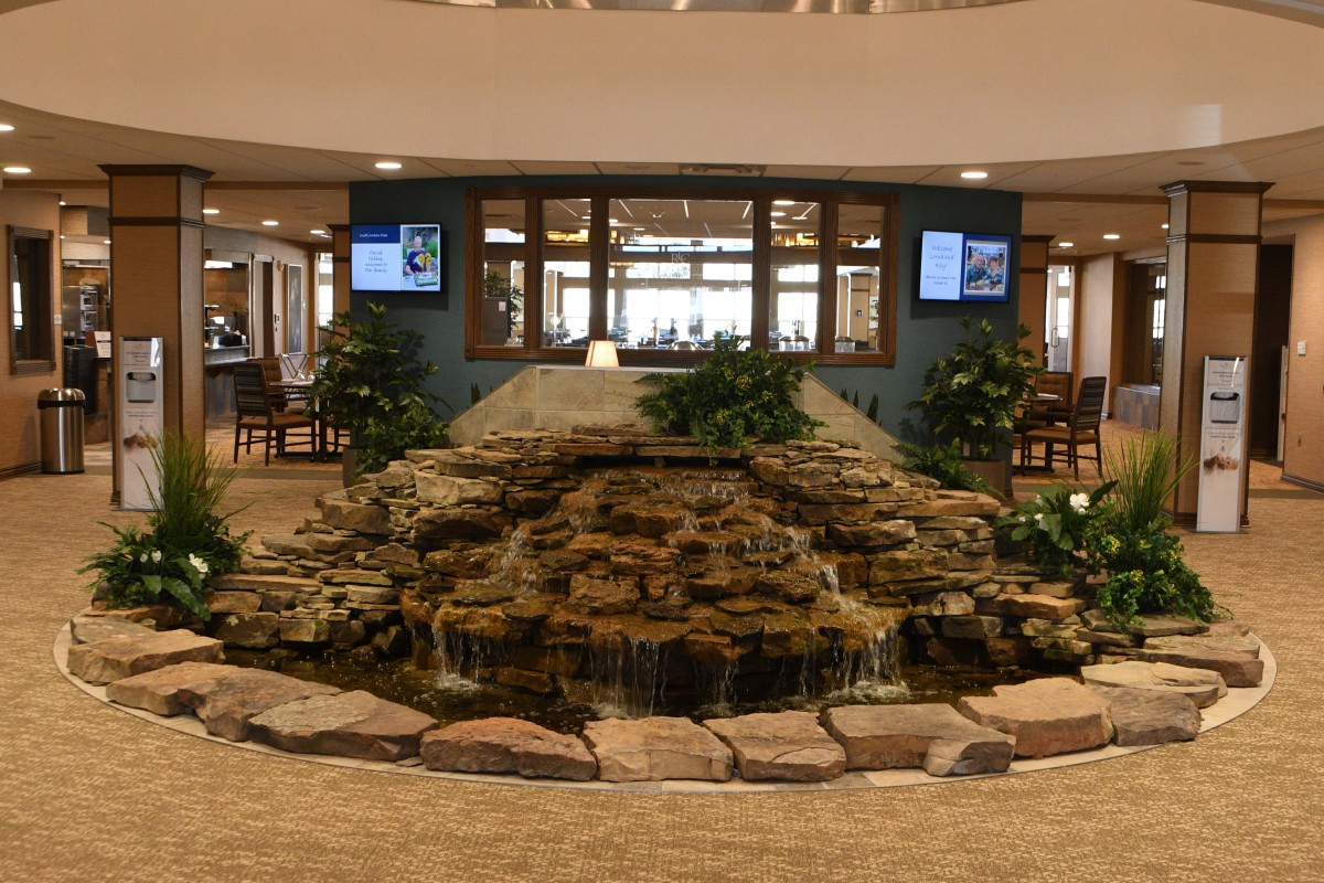 An indoor lobby featuring a central stone fountain surrounded by plants and decorative elements.