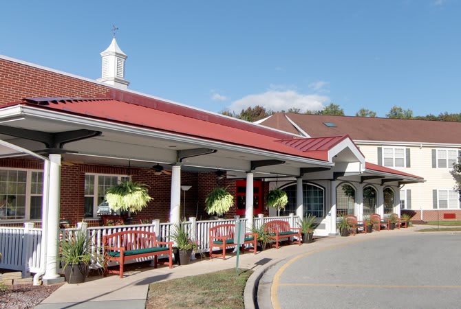 A building with a red roof and front porch adorned with benches and hanging plants on a sunny day.