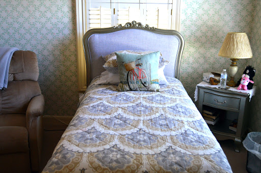 A neatly made twin bed with decorative bedding and a charming pillow, set against a patterned wallpaper in a cozy bedroom.