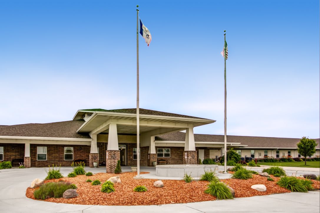 A building entrance with landscaped surroundings and flags on poles under a clear blue sky.