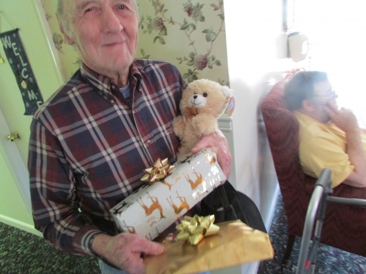 An elderly man smiling while holding wrapped gifts and a teddy bear.