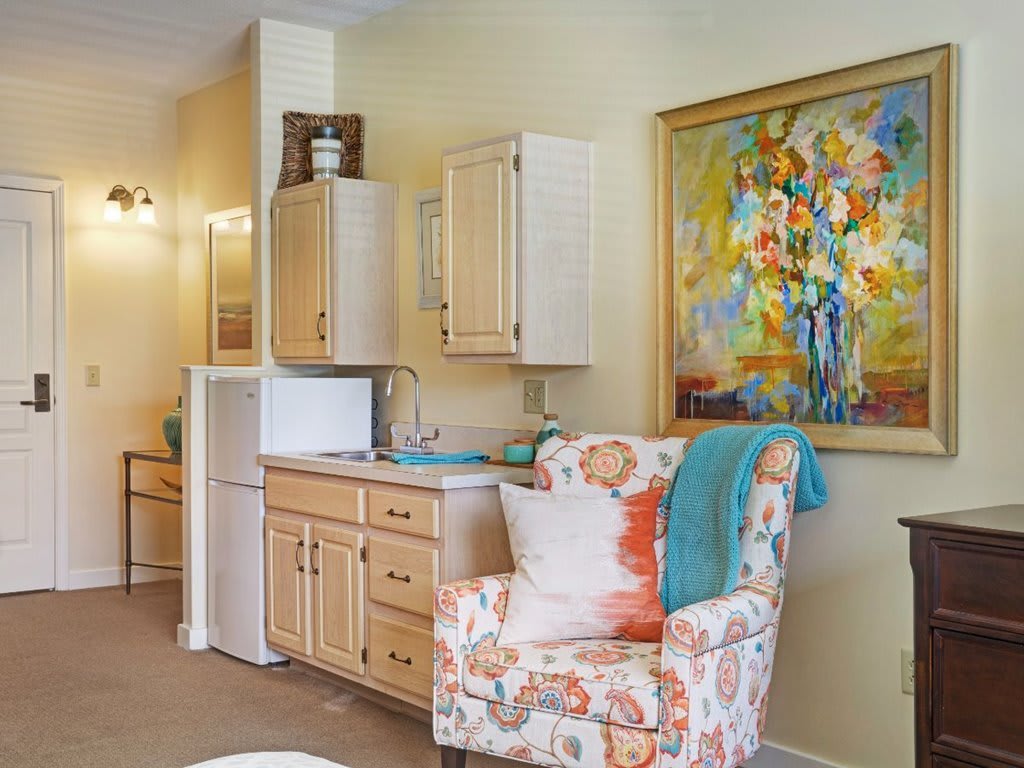 A cozy living space featuring a kitchenette, an armchair with floral patterns, and a vibrant abstract painting on the wall.