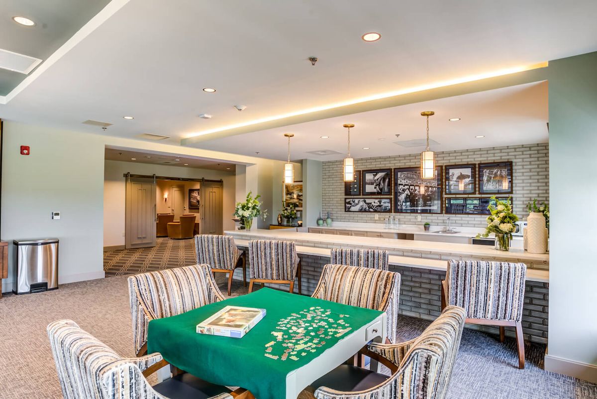 A modern and well-lit recreation room featuring a table with chairs and a kitchenette area.