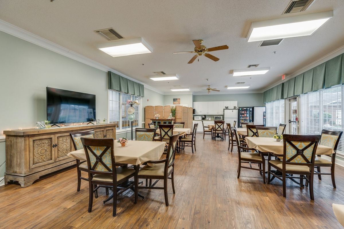 A spacious and well-lit dining room with wooden tables and chairs, featuring a large television and ceiling fan.
