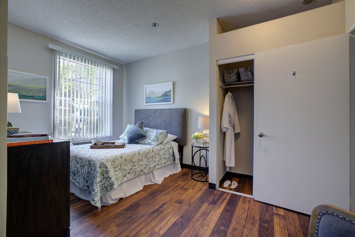 A cozy bedroom with a neatly made bed, open closet, and large window allowing natural light.
