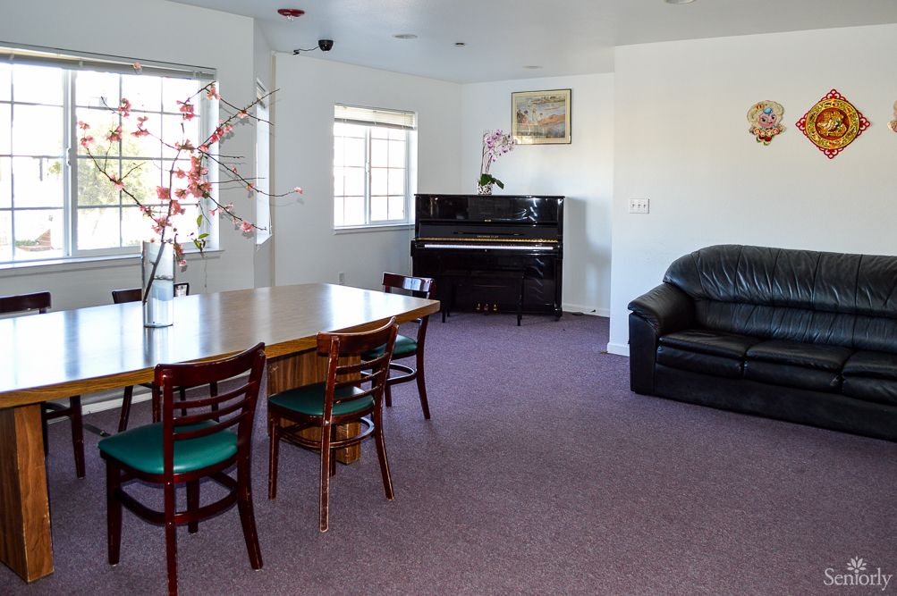 A cozy living room featuring a piano, dining table with chairs, a couch, and large windows.