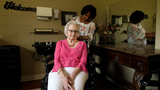An elderly woman in a wheelchair smiles as a caregiver styles her hair in a cozy indoor setting.