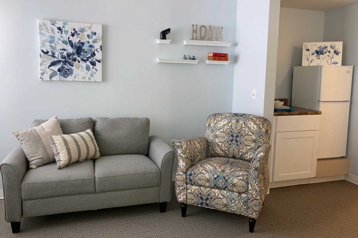 A cozy living room with a gray sofa and patterned armchair, featuring wall art and decorative shelves.