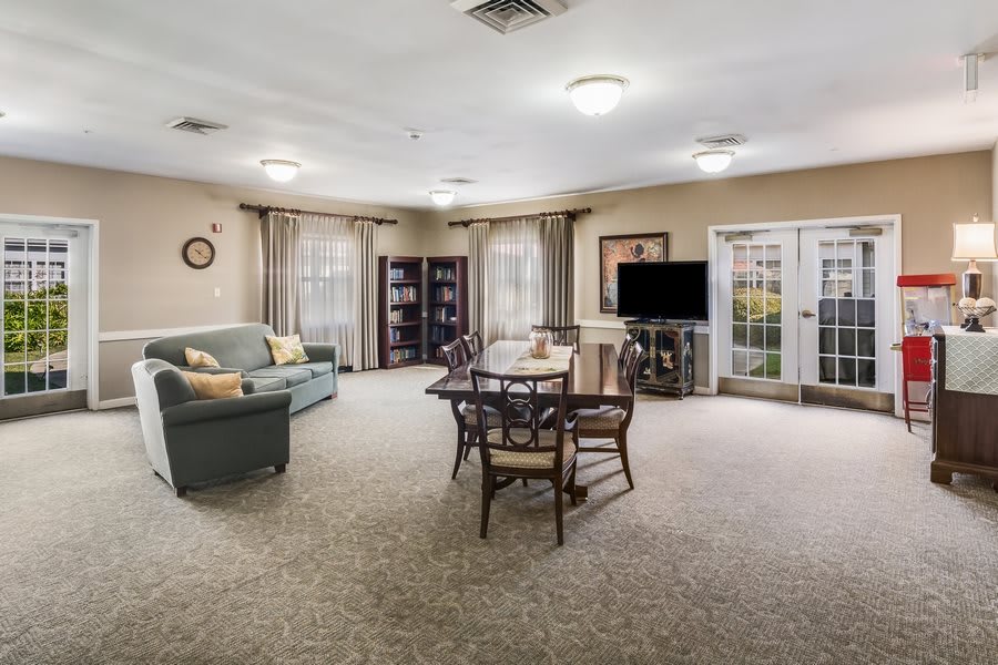 A spacious living room with a cozy sofa, dining table, bookshelf, and television, featuring carpeted flooring and ample natural light.