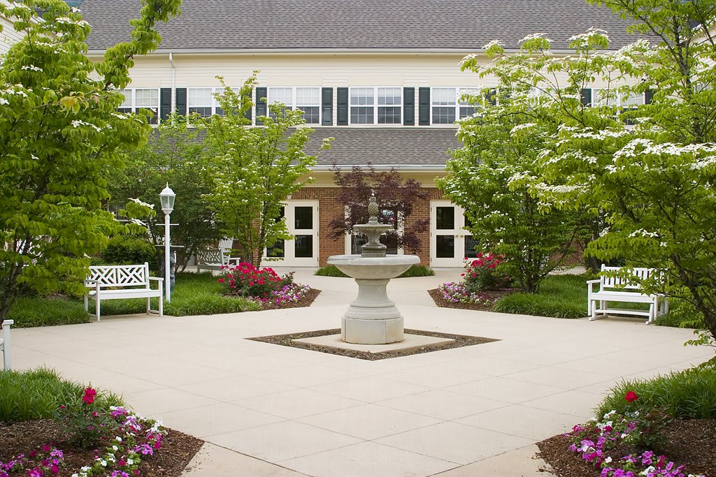 A tranquil courtyard featuring a central fountain, surrounded by benches, trees, and colorful flowers in front of a two-story building.