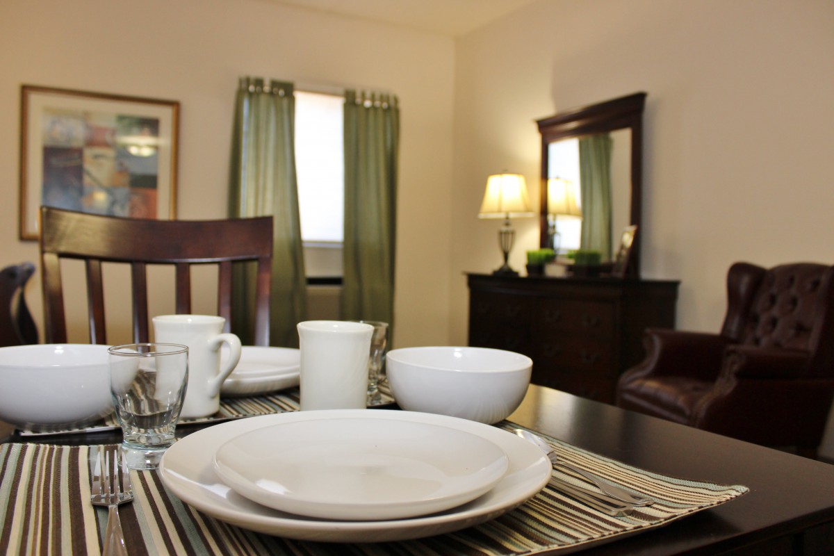 A warmly lit dining room with neatly arranged tableware on a table, featuring plates, bowls, and cups. The room includes wooden furniture and green curtains.