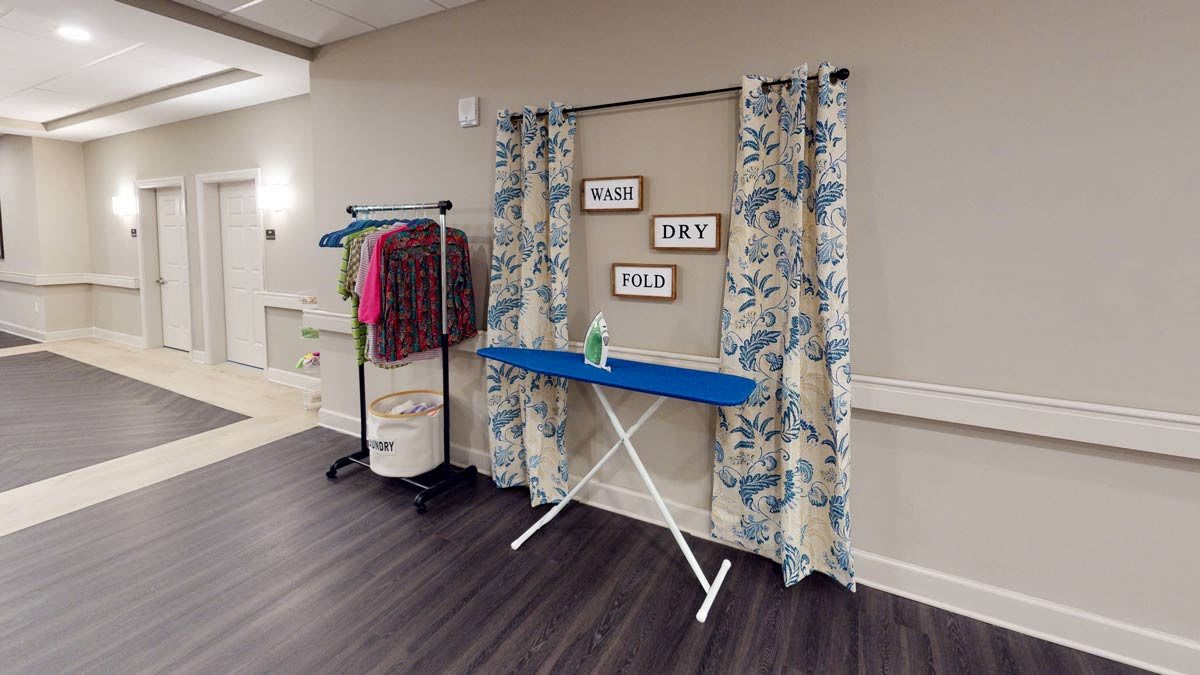 A neatly arranged laundry area featuring an ironing board, clothes rack, and decorative curtains.