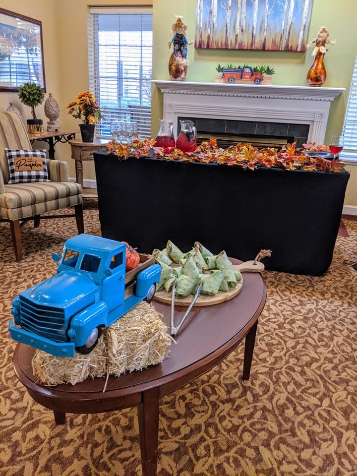An indoor autumn-themed display featuring a blue miniature truck with pumpkins on a hay bale and a fireplace adorned with fall decorations.